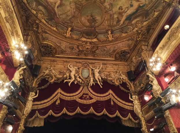 Theaterinnenraum mit roter Gardine, Kronleuchtern, Skulpturen, Lichtern und Deckenmalerei, mit einer Bühne mit ein paar Menschen davor.