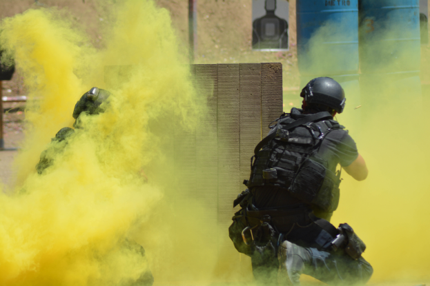 Zwei Polizisten in Schutzausrüstung mit gelbem Rauch aus dem Mund, mit Helmen und Schutzausrüstung vor einer Wand mit einer Tafel und anderen Gegenständen, mit dem Boden unten.