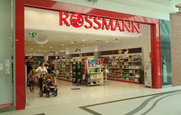 Ein Rossmann-Laden in einem Einkaufszentrum mit Deckenleuchten, Schildern, Menschen und Regalen, die mit verschiedenen Artikeln gefüllt sind, eine Person schiebt einen Kinderwagen.