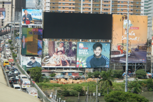 Eine belebte Stadtstraße mit starkem Verkehr, hohen Gebäuden und einem prominenten Werbeplakat, umgeben von Grünflächen und Strommasten.