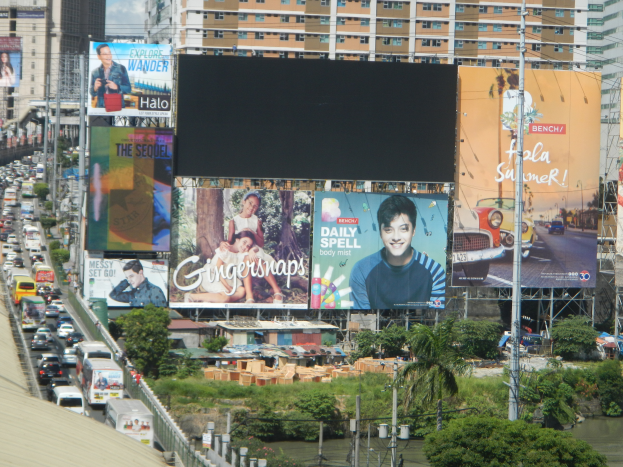 Eine belebte Stadtstraße mit starkem Verkehr, hohen Gebäuden und einem prominenten Werbeplakat, umgeben von Grünflächen und Strommasten.