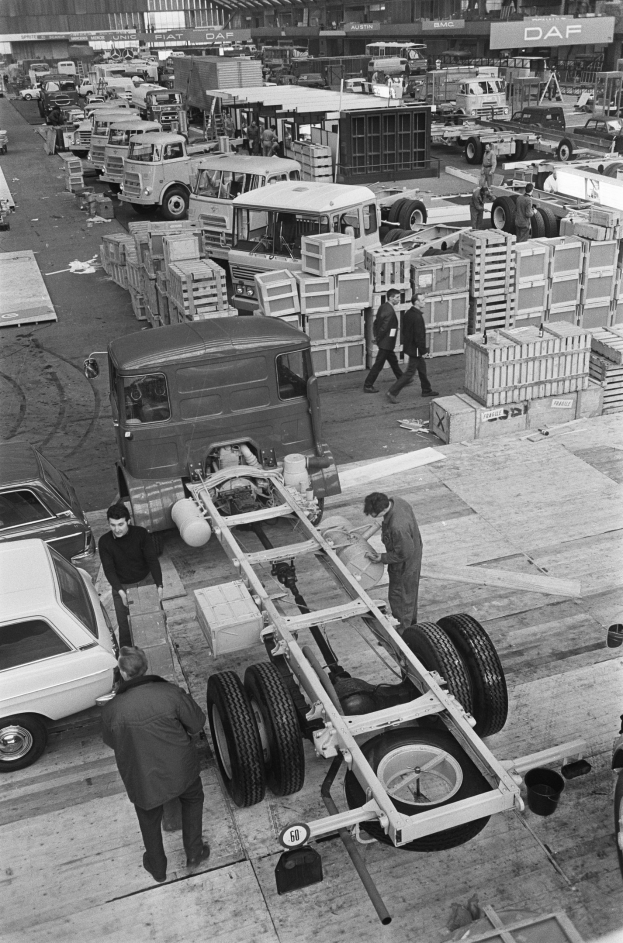 Schwarze und weiße Fotografie von Männern, die an einem Lkw in einer Fabrik arbeiten, mit Fahrzeugen, einem Rad im Vordergrund und Menschen und Kisten im Hintergrund.