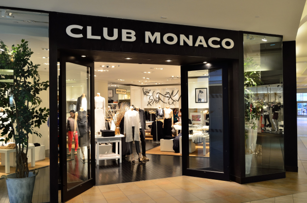 Ein Club Monaco-Laden in einem Einkaufszentrum mit Glaswänden und -türen, Schaufensterpuppen in Kleidern, Auslagetischen mit Artikeln, einer Topfpflanze und Deckenleuchten.