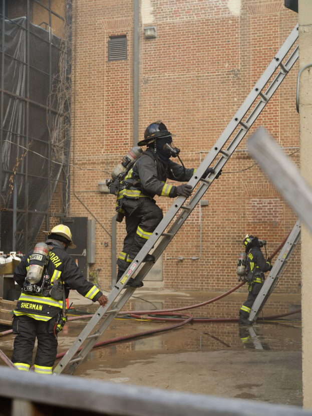 Feuerwehrleute in Helmen und Ausrüstung, die eine Leiter vor einem Backsteingebäude hochklettern, mit Rohren auf dem Boden und einer Metallstange am unteren Ende, mit einem weiteren Gebäude und einem Netz im Hintergrund.