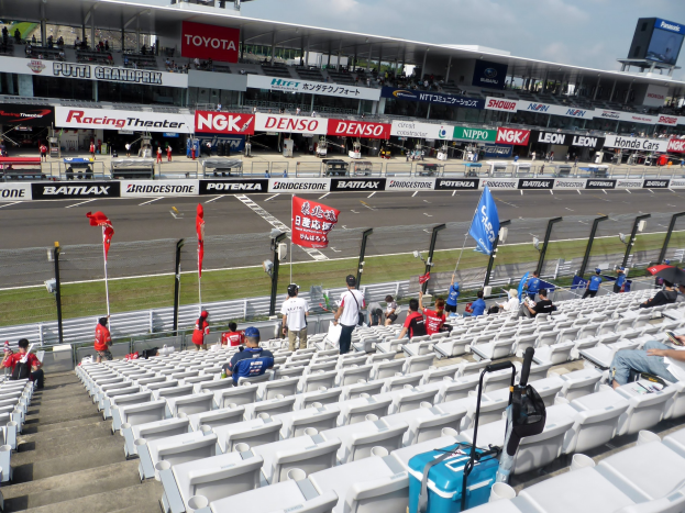 Eine Gruppe von Menschen auf Tribünen bei einer Rennstrecke mit Fahnen, Schildern, Geländern und Stufen, mit einem bewölkten Himmel über einem Stadion im Hintergrund.