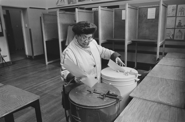 Eine Frau mit Brille hält Papiere in der Hand, während sie in einer Wahllokal mit Tischen, Bänken, Schränken, einer Tür, einem Stuhl und Plakaten im Hintergrund wählt, alles in Schwarz-Weiß.
