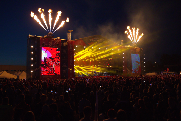 Eine große Menschenmenge versammelt sich auf einem Musikfestival mit einer hell erleuchteten Bühne, auf der Musiker spielen, umgeben von Bannern, Zelten links und Feuerwerk am dunklen Himmel.