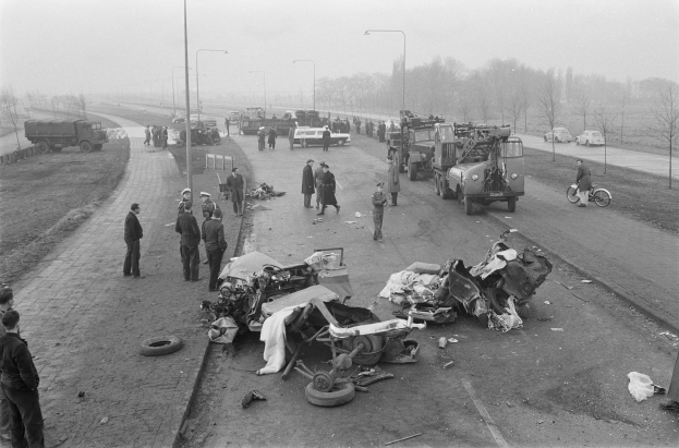 Schwarz-weiß-Szene eines Autounfalls am Straßenrand mit mehreren Fahrzeugen und einer Gruppe von Menschen drumherum, Laternen, Bäume und Himmel im Hintergrund.