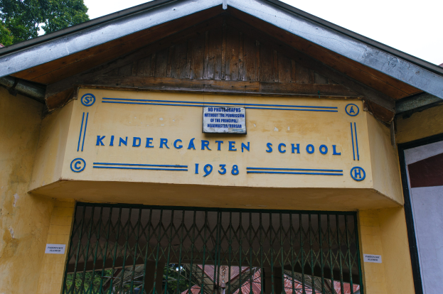 Gelbes Gebäude mit einer "Kindergarten-Schule"-Schilder, Namensschild an der Wand, ein Gitter unten, Bäume und einen klaren blauen Himmel im Hintergrund.