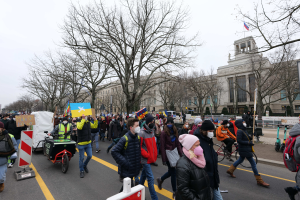 Eine große Gruppe von Menschen nimmt an einer Protestmarsch auf einer Straße in Washington, D.C. teil, einige halten Plakate und Banner, andere fahren Fahrräder und es gibt Schilder, Bäume und einen klaren blauen Himmel im Hintergrund.