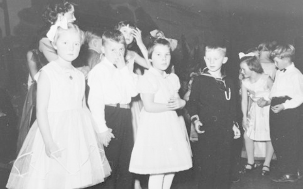 Schwarzes und weiße Bild einer Gruppe von Kindern, die auf einem Tanzboden stehen, lächeln und fröhlich aussehen, mit einer Wand im Hintergrund.