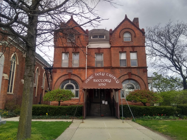 Ein großes Backsteingebäude mit einem Baum davor, umgeben von Grün, unter einem klaren blauen Himmel, mit Fenstern, einer Tür und einem Schild mit Text.