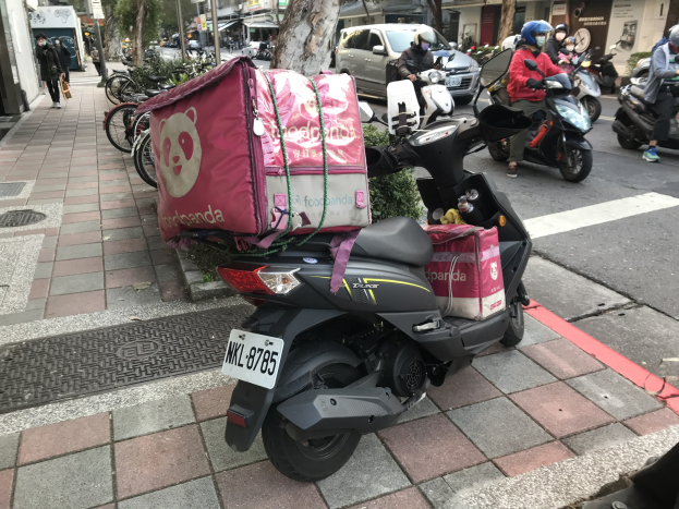 Ein Roller auf einer Straße neben einem Gebäude geparkt, mit anderen Fahrzeugen und Menschen in der Nähe und Gebäuden und Bäumen im Hintergrund.