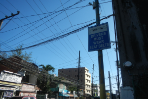 Eine Stadtstraße mit Autos, Strommasten mit Drähten, Gebäuden, Bäumen und Namensschildern, mit einem Schild an einem Mast, auf dem steht "Fahrer müssen Sitzgurte tragen" und der Himmel im Hintergrund.
