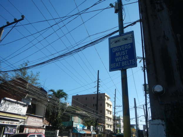 Eine Stadtstraße mit Autos, Strommasten mit Drähten, Gebäuden, Bäumen und Namensschildern, mit einem Schild an einem Mast, auf dem steht "Fahrer müssen Sitzgurte tragen" und der Himmel im Hintergrund.