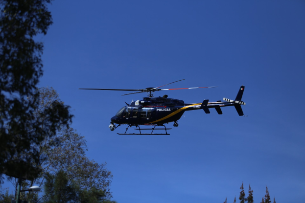 Ein Polizei-Hubschrauber fliegt durch einen blauen Himmel mit Bäumen im Vordergrund und einem Lichtmast auf der linken Seite.