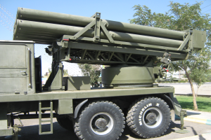 Ein Militärlastwagen mit einer Rakete fährt auf einer Straße, umgeben von Gras, Bäumen, Gebäuden und einem Maschendrahtzaun, unter einem klaren blauen Himmel.