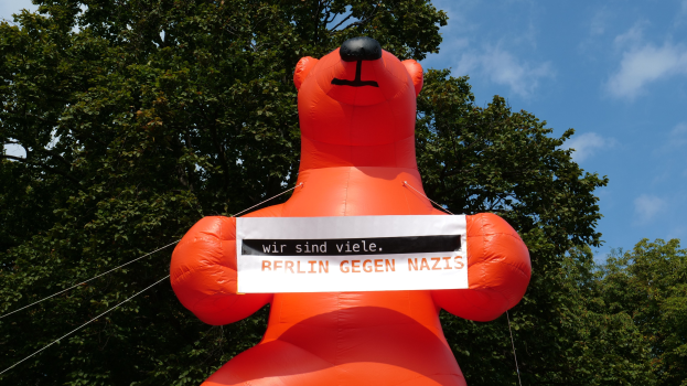 Aufblasbarer Bär mit einem Schild, auf dem "Berlin gegen Nazis" steht, vor einem Hintergrund aus Bäumen und Wolken.