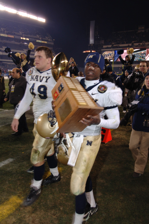 Ein Fußballspieler mit einem Pokal auf einem Feld, umgeben von einer Menge in Sportkleidung, mit Schildern, Lichtern und einem klaren blauen Himmel im Hintergrund.