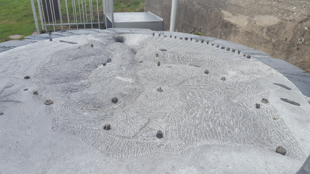 Große Betonstruktur mit kreisförmigem Design oben drauf, umgeben von einem Metallzaun, einem Pfahl, einer Wand und Gras.