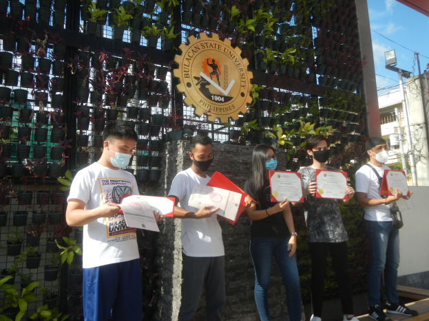 Gruppe von Menschen in Masken mit Urkunden vor einem Universitätsgebäude mit einer Pflanzenwand links und einem Bogen mit Logo und Text rechts.