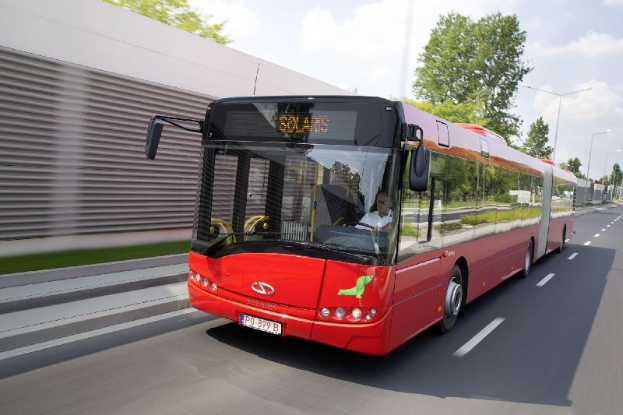 Ein roter Bus fährt auf einer Straße an einem Gebäude vorbei, mit einer Person im Inneren, neben einer Wand, Bäumen, Laternen und einem bewölkten Himmel.