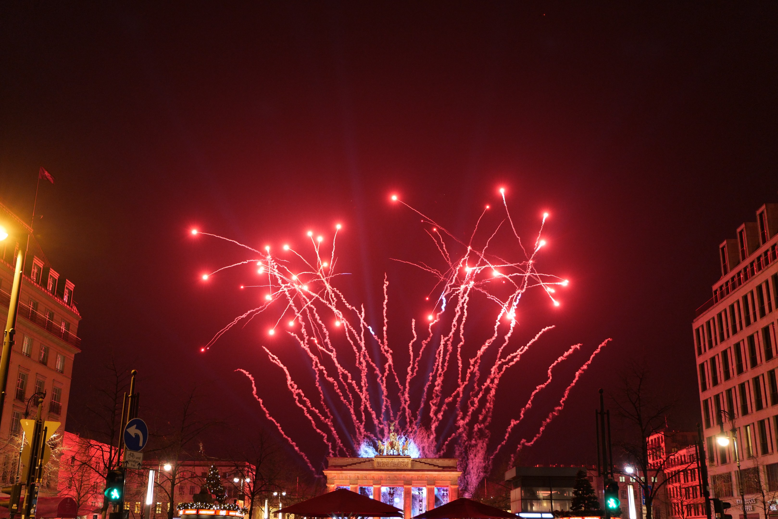 Eine nächtliche Straßenansicht in Berlin am Silvesterabend, mit Gebäuden, Bäumen, Laternenmasten, Verkehrszeichen, Zeltplanen, Menschen und einem prächtigen Feuerwerk am Himmel.