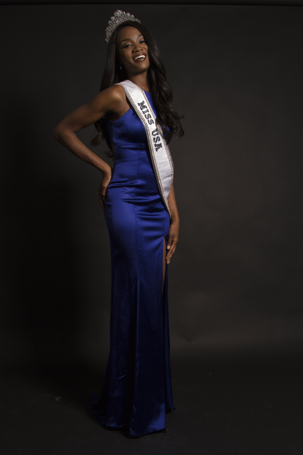 Eine Frau in einem blauen Kleid mit einer Tiara und einem Band mit der Aufschrift "Miss USA 2014" vor einem dunklen Hintergrund.