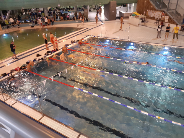 Großes Hallenbad mit schwimmenden, stehenden und sitzenden Menschen, mit Treppen, Geländern und einer Wand im Hintergrund.