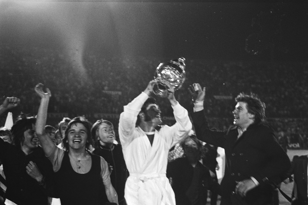 Eine Gruppe von Menschen feiert mit einem Pokal, wobei die zentrale Figur ihn in einem hell erleuchteten, schwarz-weißen Setting hochhält, während andere jubeln.