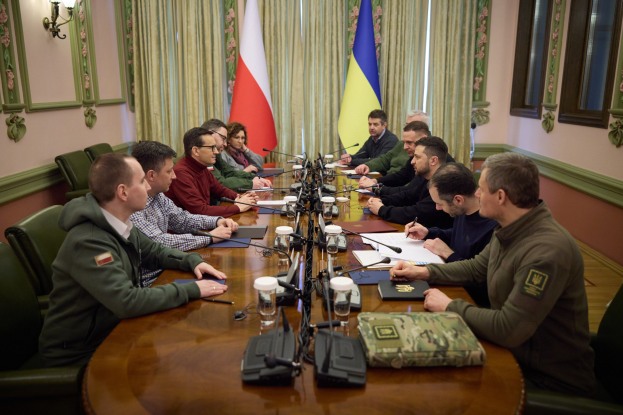 Eine Gruppe von Männern in einer offiziellen Umgebung, die um einen Holztisch mit Mikrofonen und Dokumenten sitzen und diskutieren, mit Flaggen und Lichtern im Hintergrund.