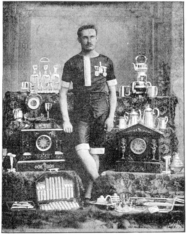 Schwarzes und weißes Foto eines Mannes in einem Fußballtrikot, der vor einem Tisch mit Pokale und Trophäen steht, beschriftet mit "1909-1910 WM-Sieger."