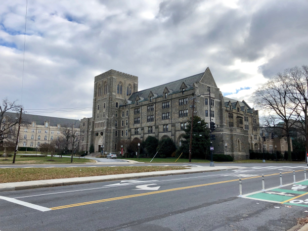 Großes Gebäude mit zahlreichen Fenstern, als der neue Campus der University of Pennsylvania bezeichnet, umgeben von Bäumen, Straßenlaternen, Verkehrsschildern und Fahrzeugen unter einem bewölkten Himmel.