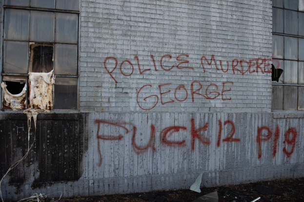 Ein Gebäude mit Graffiti, das "Polizei ermordet George Fuck 12 P19" auf der Seite trägt, mit Fenstern und verschiedenen Gegenständen davor.