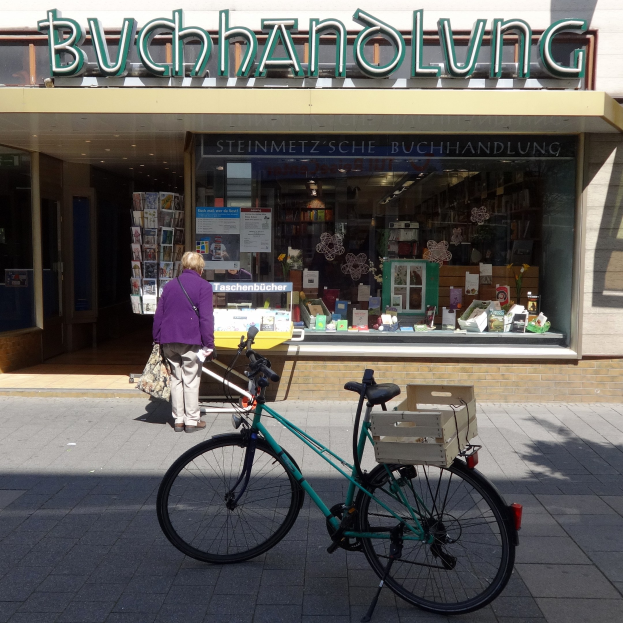 Eine Frau steht neben einem geparkten Fahrrad vor einem Laden, mit einem Fahrradrad im Vordergrund und durch das Schaufenster sichtbaren Büchern.