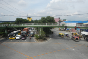 Eine belebte städtische Straße mit Autos, Bussen, Fußgängern auf einem Gehweg, einer Brücke mit Geländern, Verkehrsampeln, Bäumen, Gebäuden, Strommasten mit Drähten und einem bewölkten Himmel.