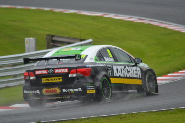 Ein Auto mit Text an der Seite auf einer regennassen Rennstrecke, umgeben von Gras, mit einem Geländer und einer hölzernen Struktur im Hintergrund.