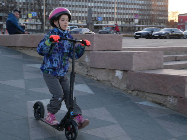 Junger Junge mit Helm und Handschuhen auf einem Roller die Straße entlang mit Treppen, Fahrzeugen, Menschen, Bäumen, Polen, Brettern, Gebäuden und einem klaren blauen Himmel im Hintergrund.