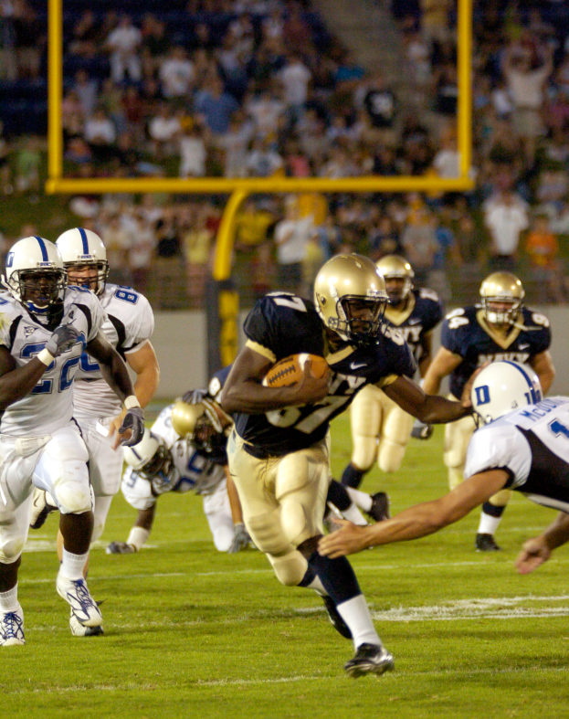 Ein Füßballspieler in einem Sportdress und Helm rennt mit dem Ball, verfolgt von einer Gruppe ähnlich gekleideter Spieler, mit unscharfen Menschen, Pfählen und Gegenständen im Hintergrund und Text unten im Bild.