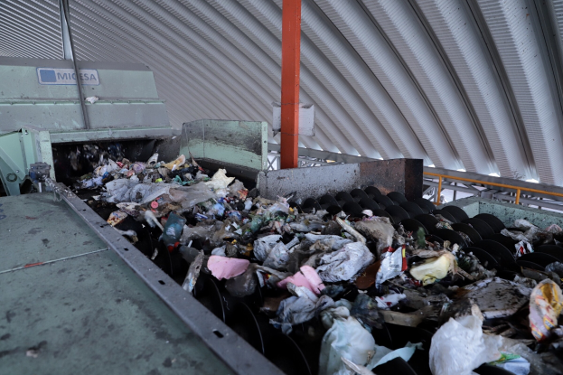 Großer Müllhaufen auf einem Förderband in einer Recyclinganlage mit Maschinen und einem Metalldach.