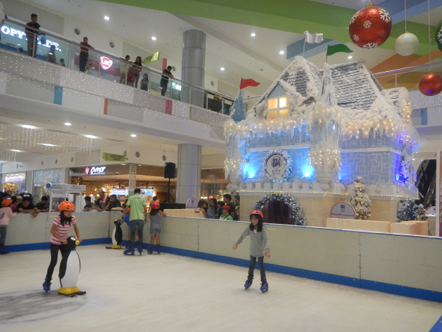 Eine Gruppe von Menschen, die auf einer Eisbahn in einem Einkaufszentrum Schlittschuh laufen, umgeben von einem hausähnlichen Aufbau, der mit Lichtern und festlichen Dekorationen geschmückt ist, mit Zuschauern, Geländern, Säulen, Texttafeln und Deckenleuchten, die eine festliche Atmosphäre schaffen.