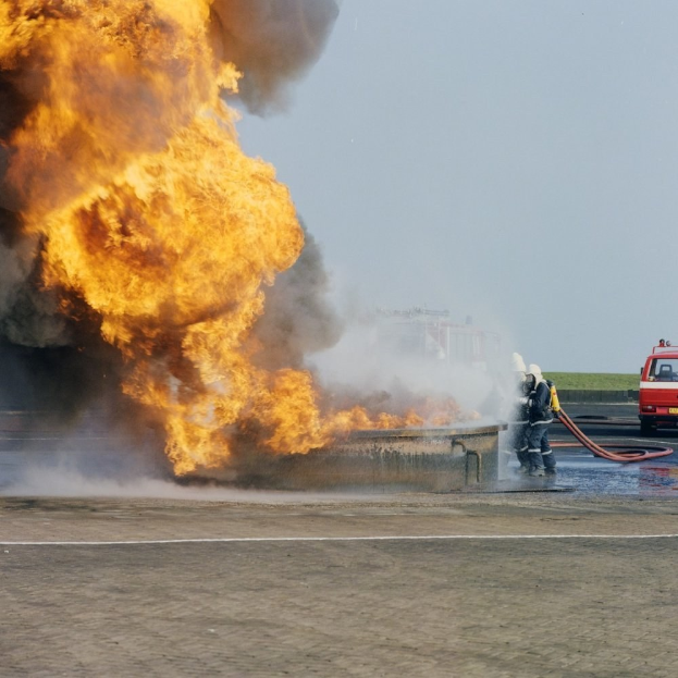 Feuerwehr-Lkw steht in Flammen am Straßenrand mit zwei Helmet tragenden Individuen, die Rohre halten, ein Fahrzeug im Hintergrund und der Himmel sichtbar.