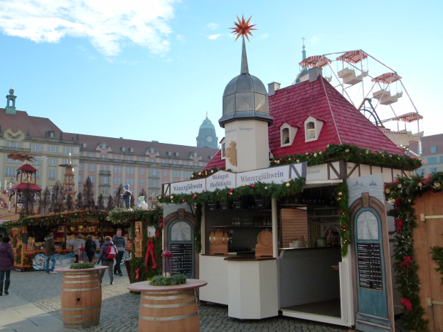 Ein geschäftiger Weihnachtsmarkt in Nürnberg, Deutschland, mit Menschen um geschmückte Stände, festliche Lichter, Schmuck, einem Riesenrad im Hintergrund, Gebäuden mit Fenstern und einem bewölkten Himmel.