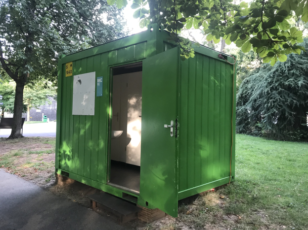 Eine grünne tragbare Toilette mit einer Tür und einer angebrachten Platte steht in einem Park, umgeben von Bäumen und einer Bank auf der linken Seite, mit dem Himmel im Hintergrund.