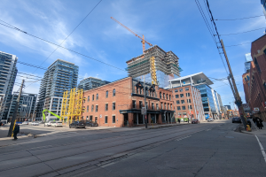 Eine belebte Stadtstraße in Toronto mit Fahrzeugen, Fußgängern, Gebäuden und einer Baustelle mit einem Kran im Hintergrund unter einem bewölkten Himmel.