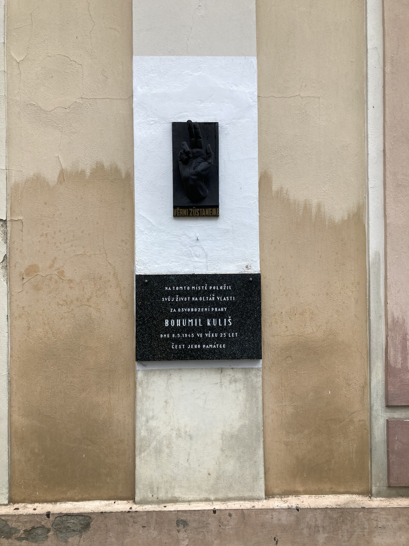 Schwarze Tafel an einem Gebäude mit einer darauf ruhenden Hand, beschriftet mit Text, der Holocaust-Opfer gedenkt.