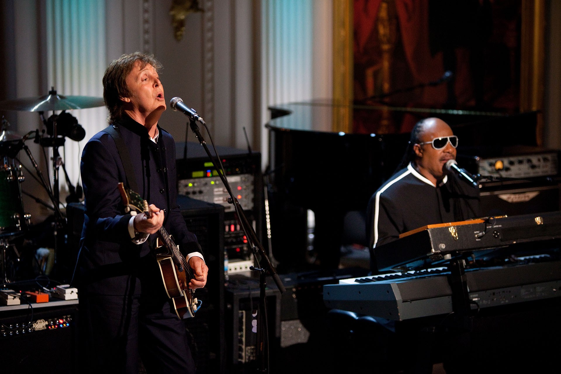 Paul McCartney spielt Gitarre und singt in ein Mikrofon, während Stevie Wonder, der eine Brille trägt, am Piano auf der Bühne der Grammys spielt, mit Instrumenten und Dekoration im Hintergrund.