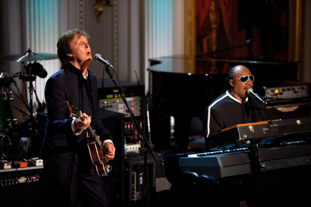 Paul McCartney spielt Gitarre und singt in ein Mikrofon, während Stevie Wonder, der eine Brille trägt, am Piano auf der Bühne der Grammys spielt, mit Instrumenten und Dekoration im Hintergrund.