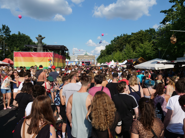 Eine große Menschenmenge, die eine Straße mit Zelten, Bäumen, Pfählen, Lichtern und einer Statue gesäumt entlanggeht, mit Gebäuden und einem bewölkten Himmel mit Ballons im Hintergrund beim Berlin Pride Festival.