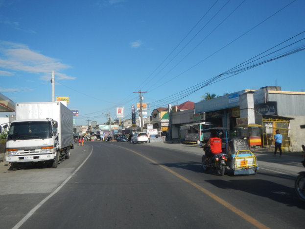 Eine Stadtstraße mit einem Lkw, der an Gebäuden, Strommasten mit Drähten, Schildern, Bäumen und einem bewölkten Himmel vorbeifährt.